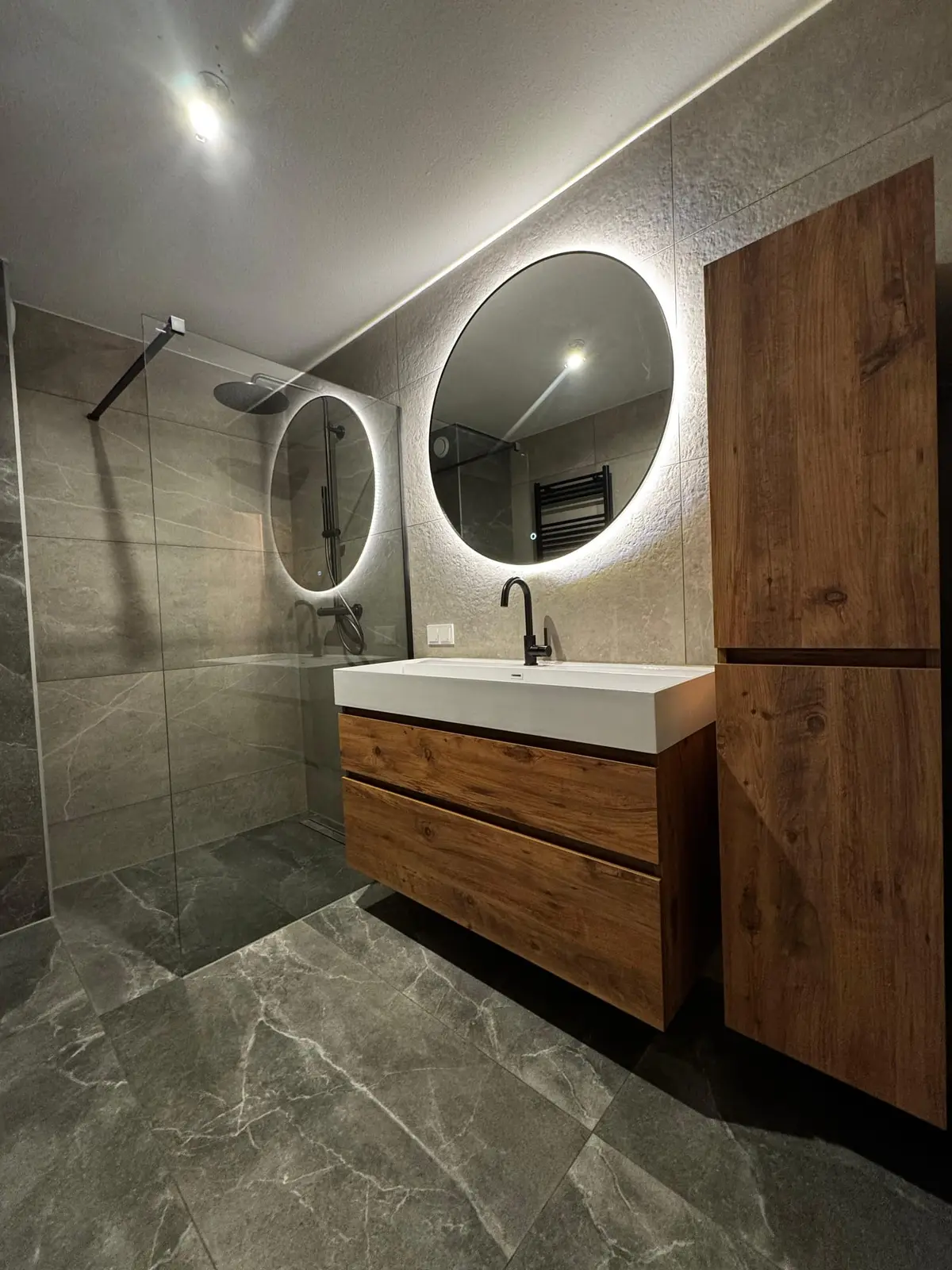 Badkamer met inloopdouche, houten wastafelmeubel en ronde verlichte spiegels